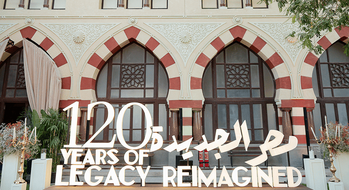 Large white sign reads '120 Years of Legacy Reimagined' in English and Arabic in front of a historic building with arched windows and red-and-white striped stonework.