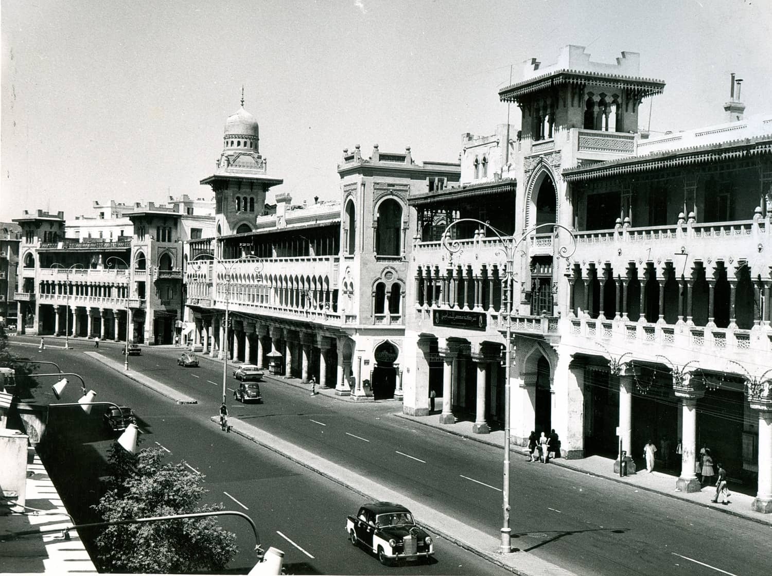 Black and white photo of an elegant, historic arcade with arches and domed towers, overlooking a street with vintage cars and pedestrians.