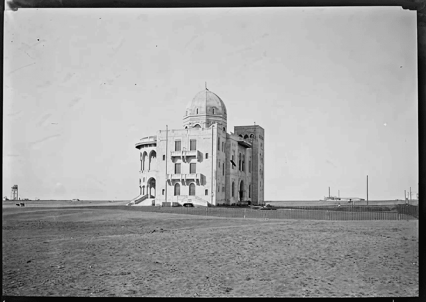 A historic building with a dome, featuring arched windows and a sandy foreground, set against a clear sky and distant coastline.