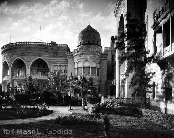 Black and white photo of an ornate, historic palace with a domed tower, surrounded by lush gardens and palm trees.