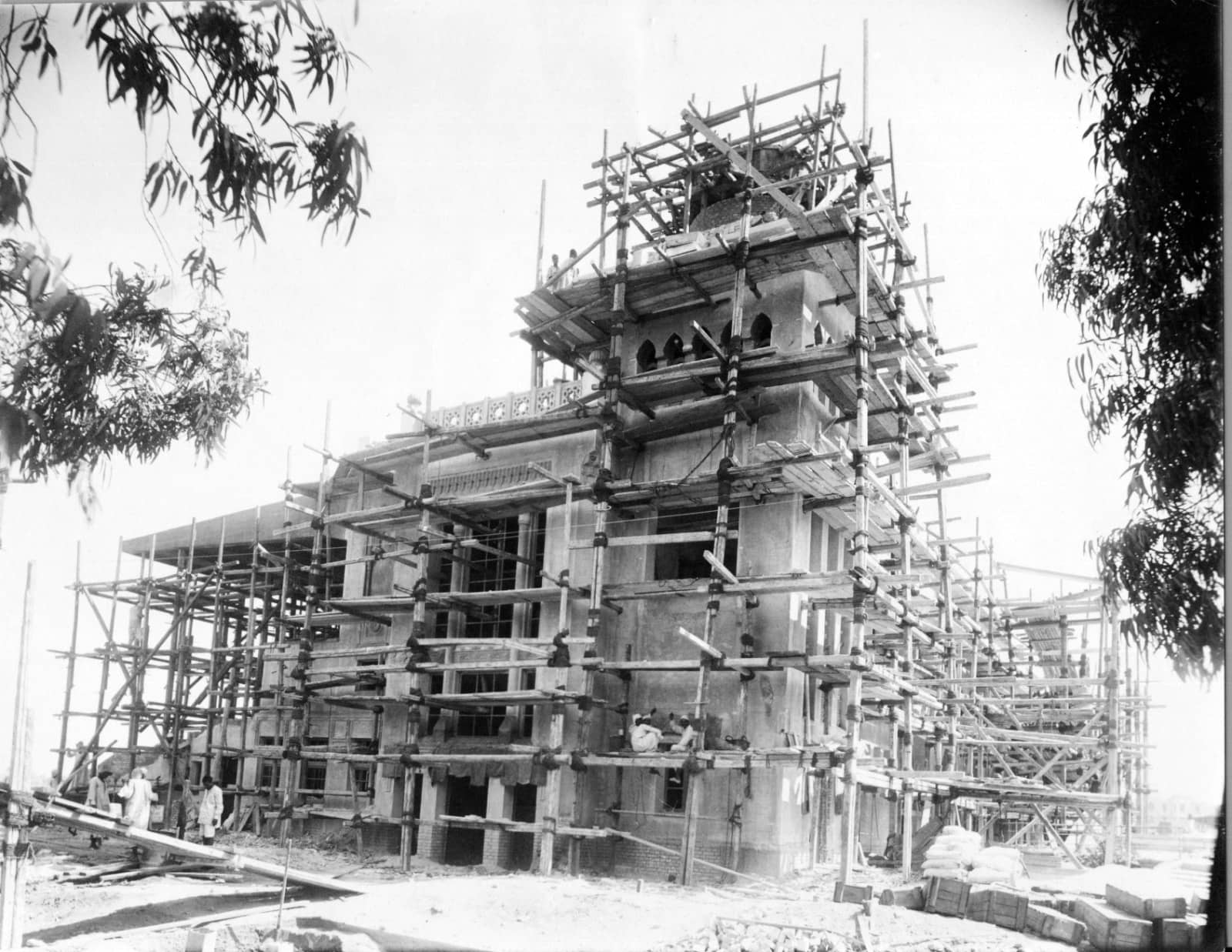 A black and white image of a building under construction surrounded by scaffolding, with a clear sky and a tree in the foreground. 