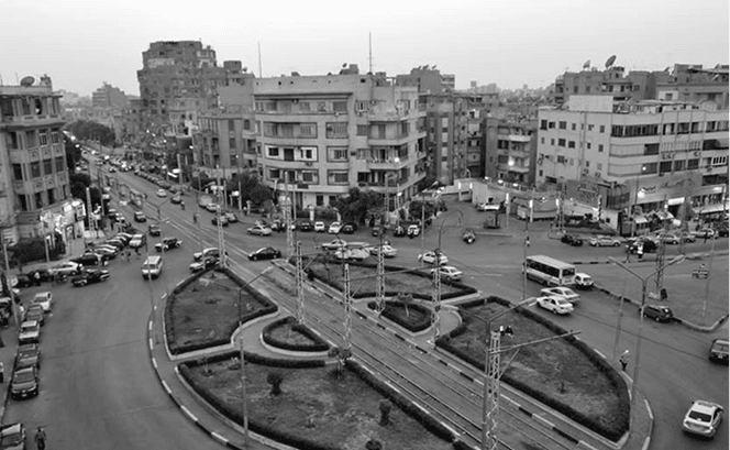 A black and white image of a street featuring cars and buildings, with a clear sky above, including an intersection in an urban area.