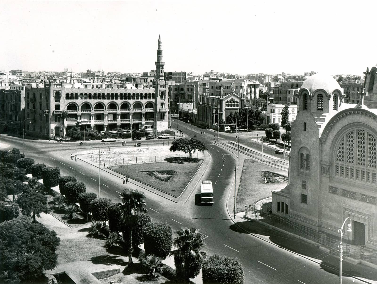 A black and white photo depicting a city scene with a road lined by buildings and trees, under a cloudy sky. Cars are parked along the street near a government building.