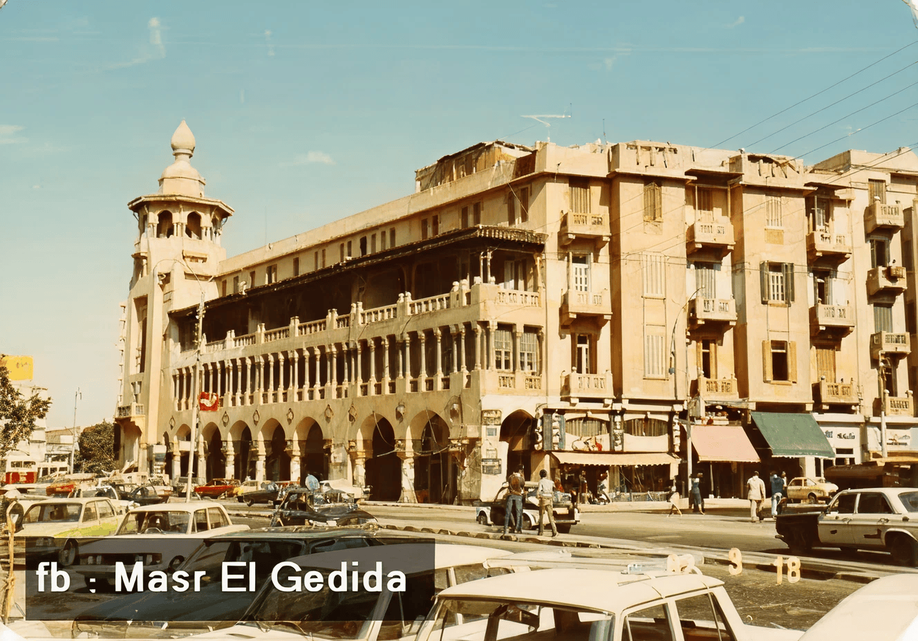 18)	Street view of a distinctive building with arched colonnades and a tower, lined with cars and shops under a clear sky.