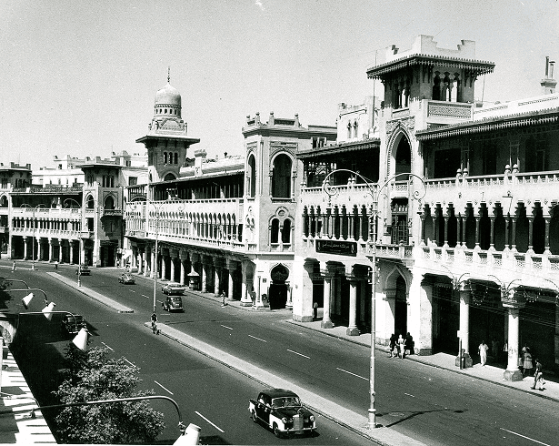 A black-and-white photograph of a historic street scene featuring ornate colonial buildings, a wide road, and vintage cars.