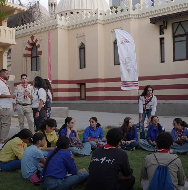 Large white sign reads '120 Years of Legacy Reimagined' in English and Arabic in front of a historic building with arched windows and red-and-white striped stonework.