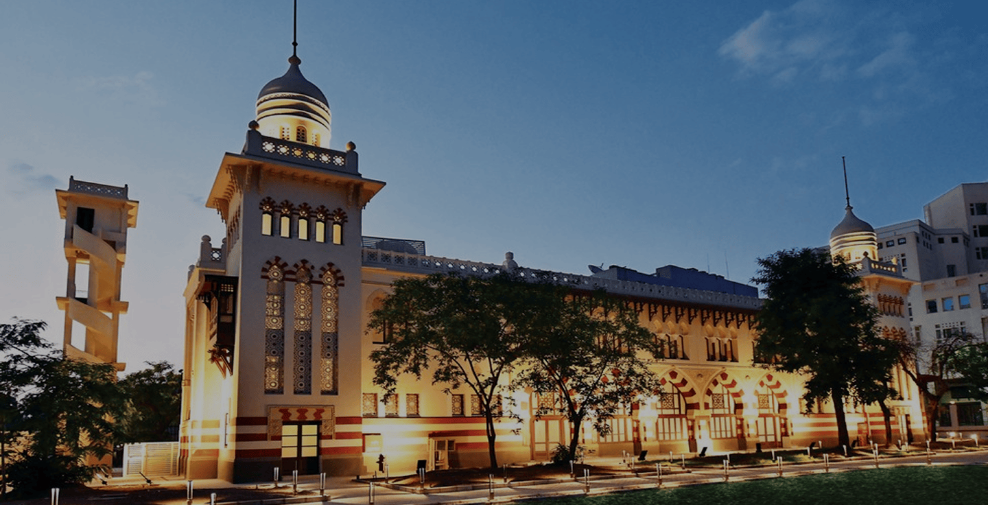 Black and white photo of an elegant, historic arcade with arches and domed towers, overlooking a street with vintage cars and pedestrians.