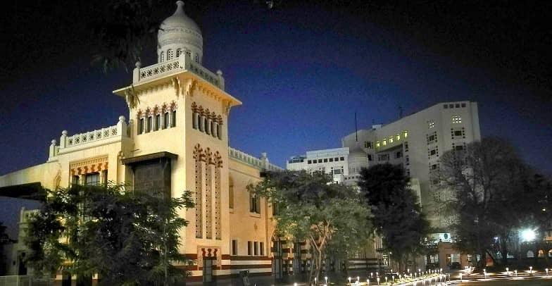 Large white sign reads '120 Years of Legacy Reimagined' in English and Arabic in front of a historic building with arched windows and red-and-white striped stonework.